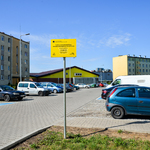Parking dla mieszkańców ulic Zagórnej i Jarzębinowej z zaparkowanymi samochodami, w tle widoczny sklep Biedronka, na pierwszym planie żółta tabliczka informująca o budowie parkingu w ramach Budżetu Obywatelskiego 2018