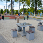 Soliki do gry w szachy, z tyłu ludzie ćwiczą na siłowni zewnętrznej oraz dzieci skaczą na trampolinach.