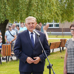 Zdjęcie terenu zielonego przed szkołą. Na pierwszym planie prezydent przemawia do mikrofonu, obok stoi dyrektorka szkoły. Za nimi, w oddali kilkoro uczniów. W tle drzewo i budynek szkoły.