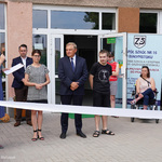 Na pierwszym planie rozciągnięta biała szarfa do przecięcia. Prezydent Białegostoku, jego zastępca, dyrektorka szkoły, kilku nauczycieli i uczniów stoi za szarfą, przed budynkiem szkoły.