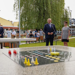 Zdjęcie terenu zielonego przed szkołą. Na pierwszym planie stół do gry w chińczyka, dalej prezydent przemawia do mikrofonu, obok stoi dyrektorka szkoły i rzeczniczka prasowa miasta. Za nimi i obok nich, w oddali kilkoro uczniów. W tle drzewo i budynek szkoły.