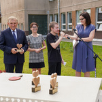 Teren otwarty przed szkołą. Na pierwszym planie stół do pingponga, na którym stoją statuetki w kształcie serc, za nim prezydent Białegostoku trzymający statuetkę, dyrektorka szkoły, rzeczniczka urzędu miejskiego oraz uczeń mówiący do mikrofonu. W tle budynek szkoły.