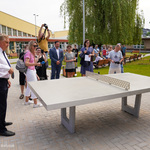 Teren otwarty przed szkołą. Na pierwszym planie stół do pingponga przy którym prezydent Białegostoku oraz uczeń grają w grę. Za nimi uczniowie, nauczyciele i dziennikarze robiący zdjęcia. W tle budynek szkoły.