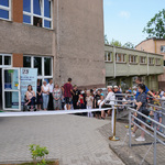 Kilkadziesiąt osób stojących przed budynkiem szkoły. Na pierwszym planie rozciągnięta biała szarfa do przecięcia.