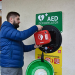 Mężczyzna wyjmuje z zamontowanej na ścianie skrzynki defibrylator AED.