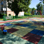 Plac zabaw otoczony zielonym płotem. Kolorowa nawierzchnia z poliuretanu. Drzewo rzuca cień. W oddali blok.
