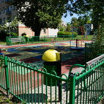 Plac zabaw otoczony zielonym płotem. Drzewo rzuca cień. W oddali blok.