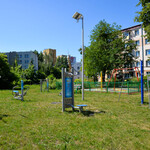 Urządzenia do ćwiczeń na świeżym powietrzu, trawa. W oddali plac zabaw, drzewa, blok. Bezchmurne niebo.