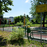 Plac zabaw otoczony zielonym płotem. Żółta tablica informacyjna. Wysokie drzewa, bezchmurne niebo.