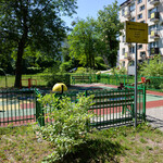 Plac zabaw otoczony zielonym płotem. Drzewo rzuca cień. W oddali blok.