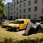 Parking przed blokiem. Kilka samochodów i motocykl przykryty pokrowcem.