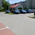 Parking wyłożony kostką. Kilkanaście samochodów. W oddali niewysoki budynek, w tle bloki.