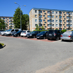 Niebieskie niebo. Kilka samochodów na parkingu. W tle drzewa i bloki.