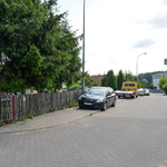 Widok na skrzyżowanie ulic. Nawierzchnia z kostki brukowej. Samochoy stojące na ulicy. Płot, drzewo, w oddali kilka budynków.