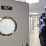 Uchylone drzwi do szatni męskiej z okrągłym okienkiem. Mężczyzna wskazuje na korytarz za drzwiami.