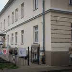 Ulica, piętrowy budynek, przy nim metalowe ogrodzenie na którym wisi biała skrzyneczka.