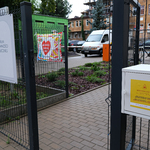 Ulica, metalowe ogrodzenie, na którym wisi biała skrzyneczka. W tle parking, samochody i budynki.