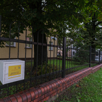 Ulica, chodnik, metalowe ogrodzenie, na którym wisi biała skrzyneczka. W tle drzewa.
