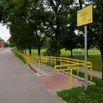 Na pierwszym planie zdjazd dla rowerów. po lewej ulica i budynki, po prawej przestrzeń parku.