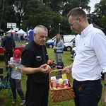 Zastępca prezydenta miasta wśród ludzi. W dłoni trzyma pleciony kosz. Rozdaje jabłka, które leżą w koszu. W tle namioty, ludzie, drzewa.