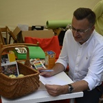 Zastępca prezydenta siedzi przy stoliku pod namiotem, w dłoni trzyma kartkę. Na stole stoi kosz z jabłkami i broszurami informacyjnymi.