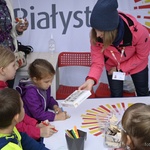 Grupa dzieci przy stoliku pod namiotem. Kobieta pochyla się na stołem. W dłoni trzyma pudełko z puzzlami.