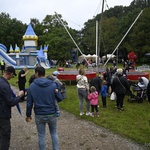 Otwarta przestrzeń parku. Grupy dzieci i dorosłych chodzą po trawniku lub stoją przed namiotami, trampolinami i dmuchanym zamkiem. W tle wysoki drzewa.