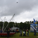 Otwarta przestrzeń parku. Grupy dzieci i dorosłych chodzą po trawniku lub stoją przed trampolinami, dmuchanym zamkiem i samochodem Straży Miejskiej. Nad nimi unosi się dron. W tle wysoki drzewa.