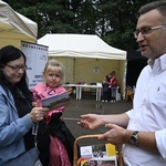 Na pierwszym tle zastępca prezydenta miasta. W dłoni trzyma pleciony kosz z broszurami informacyjnymi. Obok stoi kobieta. Na ręku trzyma dziecko, w drugiej dłoni broszurę informacyjną. W tle namioty, drzewa.