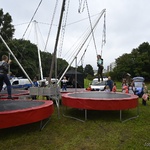 Otwarta przestrzeń w parku. Dzieci skaczą na dużych trampolinach. W tle drzewa i budynki.