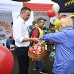 Zastępca prezydenta miasta wśród ludzi. W dłoni trzyma pleciony kosz. Rozdaje jabłka, które leżą w koszu. W tle namioty, ludzie.