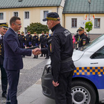 Przestrzeń miejska. Trzej mężczyźni. Dwaj podają sobie dłonie. Obok samochód. W tle ludzie, budynki.
