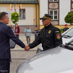 Przestrzeń miejska. Dwaj mężczyźni podający sobie dłonie. Obok samochód. W tle ludzie, budynki.