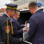 Przestrzeń miejska. Trzej mężczyźni przy samochodzie. Jeden z nich trzyma niewielkie pudełko. W tle budynki.