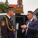 Przestrzeń miejska. Dwaj mężczyźni rozmawiają przy samochodzie. W tle kościół.