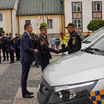 Przestrzeń miejska. Trzej mężczyźni. Dwaj podają sobie dłonie. Obok samochód. W tle ludzie, budynki.