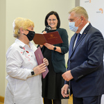 Jasne pomieszczenie. Prezydent Miasta w maseczce na twarzy stoi na przeciwko kobiety o jasnych włosach, która trzyma w dłoniach teczkę. Za nimi stoi kobieta o ciemnych włosach.