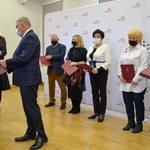 Jasne pomieszczenie. Prezydent Miasta ściska dłoń kobiety w białej bluzce. Podaje jej teczkę. Obok stoi kilka innych osób z teczkami w dłoniach.