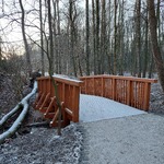 Piaszczysta ścieżka wśród drzew. Widok na drewniany mostek nad rowem.