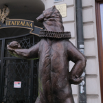 Mały metalowy miś z kryzą na szyi i i wyciągniętą do przodu łapką. W tle ściana budynku i brama z napisem akademia teatralna.