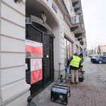 Chodnik przy budynku z dużą, metalową bramą. Dwóch mężczyzn stoi przy budynku. Obok leżą różne narzędzia.