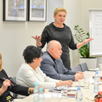 Kobieta o krótkich blond włosach stoi przy stole i mówi do zebranych gestykulując.