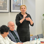 Kobieta o krótkich blond włosach stoi przy stole i mówi do zebranych gestykulując.