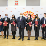 Dziewięć osób w maseczkach na twarzach pozuje do zdjęcia na ściance z logo Miasta Białystok w jasnym pomieszczeniu.