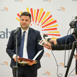 Jasne pomieszczenie. Mężczyzna w ciemnym garniturze mówi do mikrofonu na statywie i mikrofonów trzymanych  w dłoniach osób znajdujących się poza kadrem.