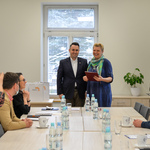 Zastępca prezydenta pozuje do zdjęcia z krótkowłosą blondynką trzymającą teczkę. Siedzący przy stole patrzą w ich kierunku.