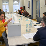 Kilka osób siedzi przy stole. Mężczyzna w żółtym swetrze wrzuca kartkę do urny.