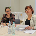 Kobieta o rudych włosach mówi do zgromadzonych. Obok niej siedzi kobieta we wzorzystej marynarce.
