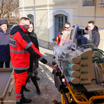 Mężczyzna w jaskrawej kurtce z napisem lekarz na plecach pokazuje zebranym inkubator.