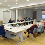Przestronne pomieszczenie, grupa kilkunastu osób siedzi przy stole ustawionym w kształt litery U na środku pomieszczenia. Na ekranie rzutnika na jednej ze ścian wyświetlana jest prezentacja. 